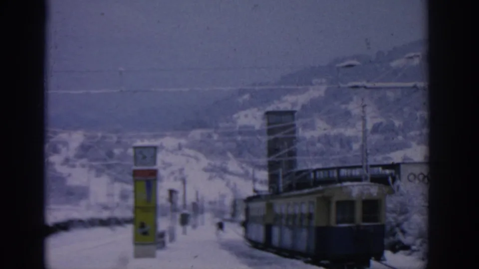 Vintage 8mm frame of a blue and yellow tram at a snowy Munich-area station, with a clock sign and mountain slope behind it.