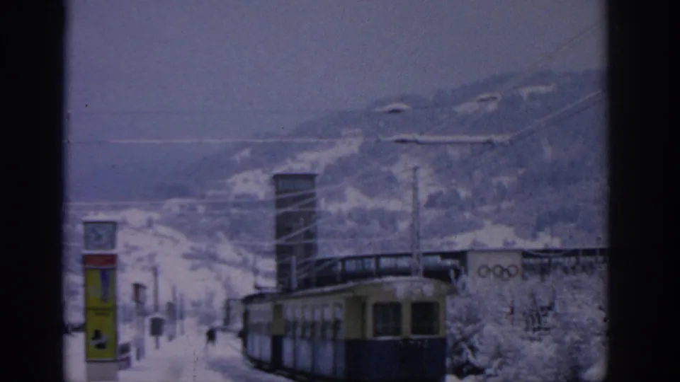 1963 Munich-area tram station view with snow, overhead wires, a clock sign, distant pedestrians, and visible Olympic-ring signage.