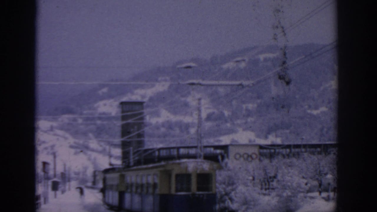Snowy rail scene with a yellow-blue tram, overhead wires, hills, and Olympic rings on a building.