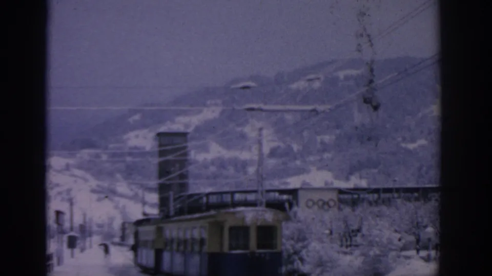 1963 Munich home-movie frame showing a yellow-blue tram below snowy hills, with overhead wires and Olympic rings visible on a nearby building.