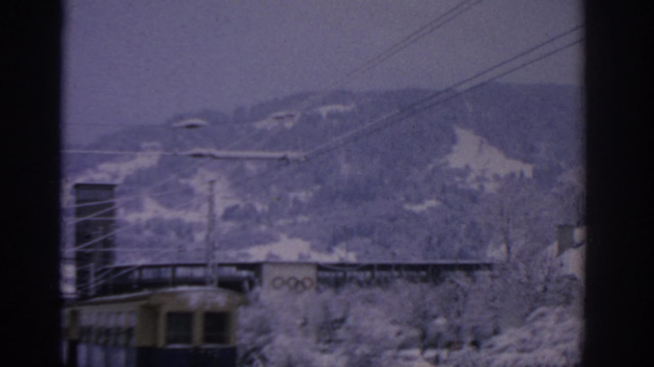 Snowy hillside view with tram wires, a rail vehicle, and Olympic rings on a building.