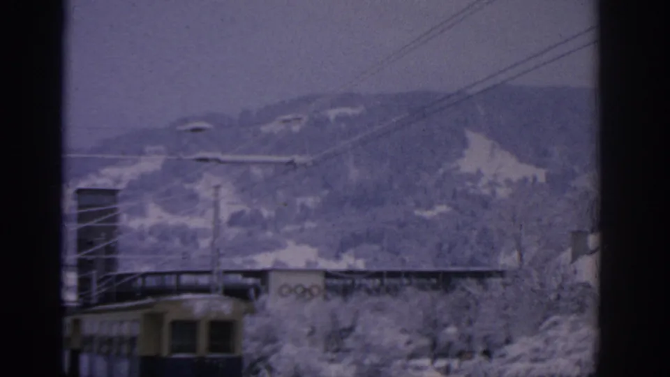 1963 Munich frame with snowy hills in the background, electric rail wires overhead, and Olympic rings on a low building near a yellow-blue tram.