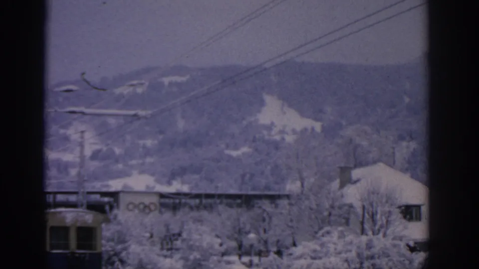 1963 Munich home-movie view of snow-covered buildings and wooded hills, with overhead rail wires and Olympic rings partly visible near the lower edge.