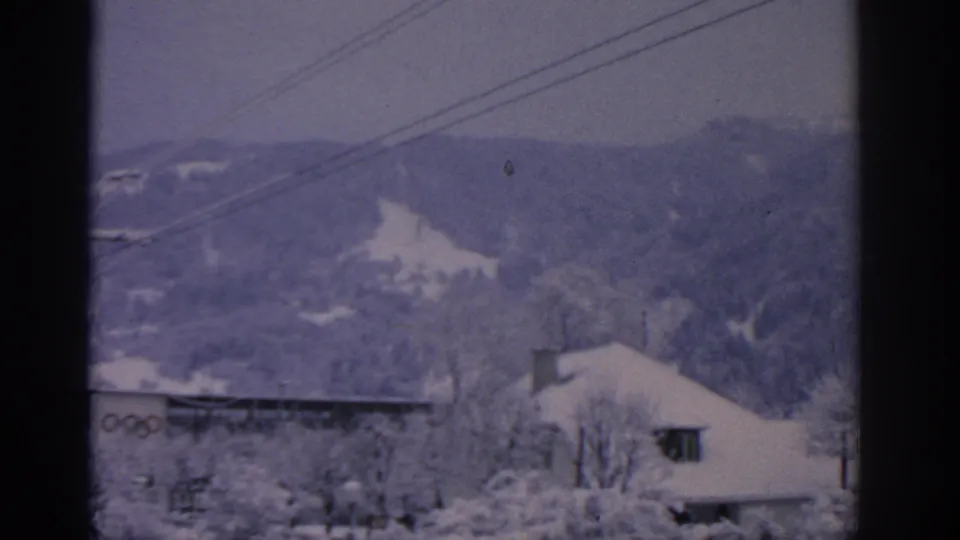 1963 Munich winter frame showing snow-covered buildings, wooded hills, overhead wires, and a partial Olympic rings sign near the lower left.