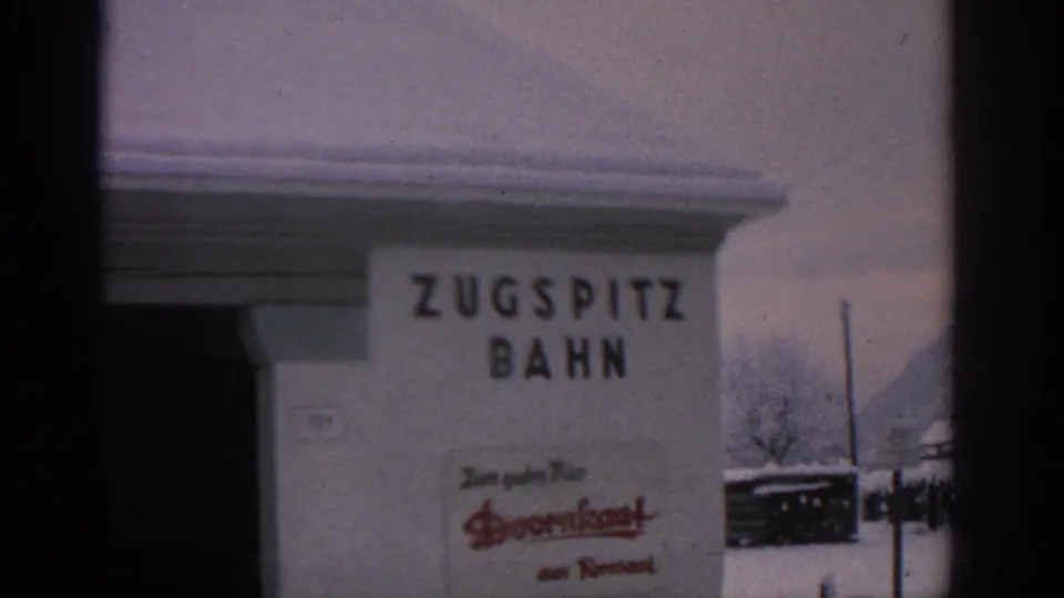 Close view of a Zugspitzbahn wall sign in 1963 Germany, with snow on the roof and a Doornkaat advertisement below.