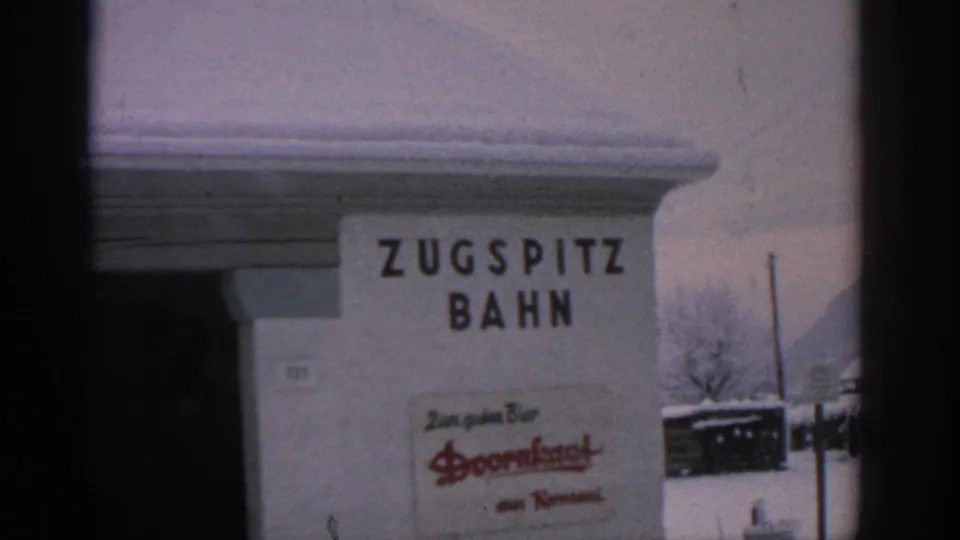 1963 8mm close-up of the Zugspitzbahn sign on a snowy German station wall, with a Doornkaat brand sign visible below.