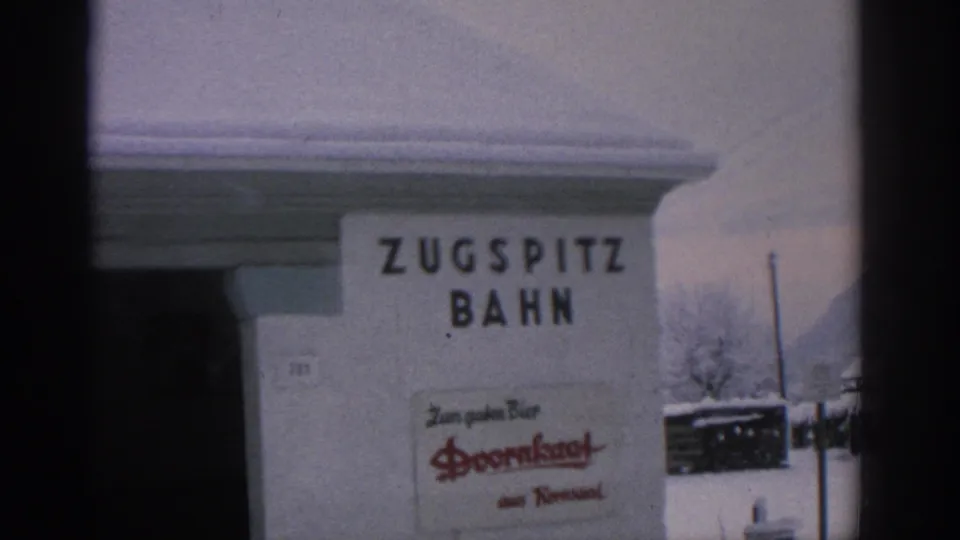 1963 German home-movie frame showing a snow-covered Zugspitz Bahn station sign with a visible Doornkaat advertisement.