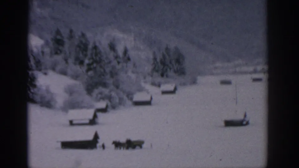 1963 German alpine winter view with small cabins scattered across a snowy slope below dark trees.