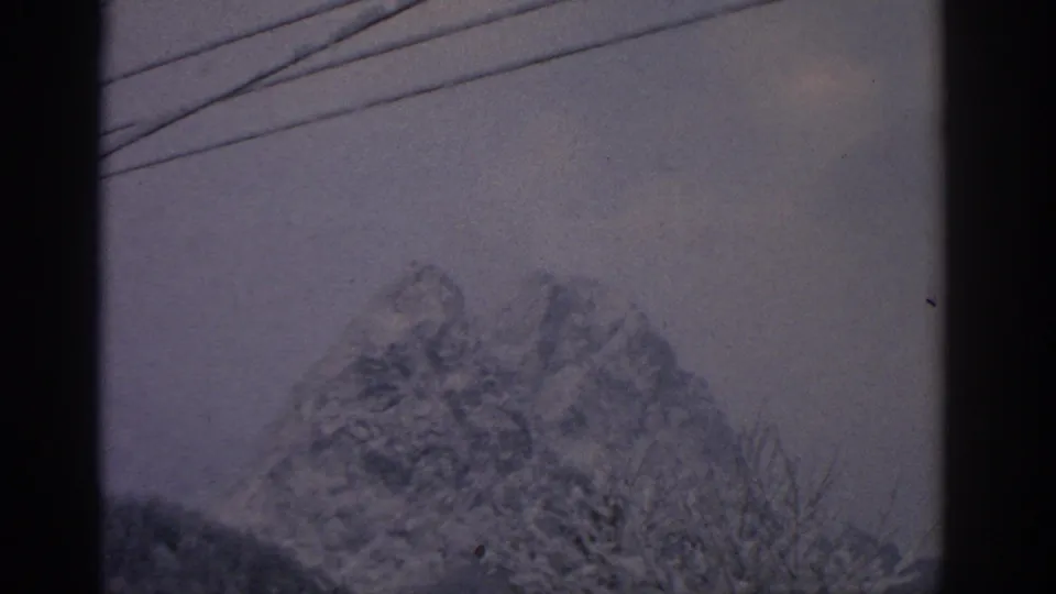 Distant snow-covered mountain near Munich in 1963 footage, with overhead wires, bare branches, and cloudy sky visible.