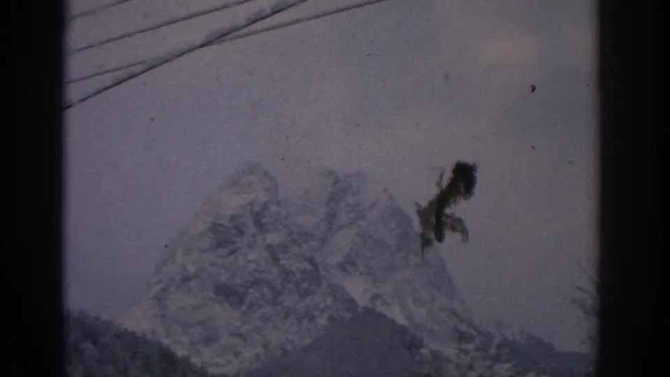 Final 1963 Munich-area mountain frame with cloudy sky, overhead wires, bare branches, and a visible dark film blemish.