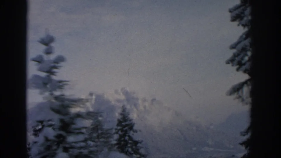 1963 8mm home-movie view near Munich showing snowy alpine peaks, dark trees, pale sky, and a hazy lower ridge.