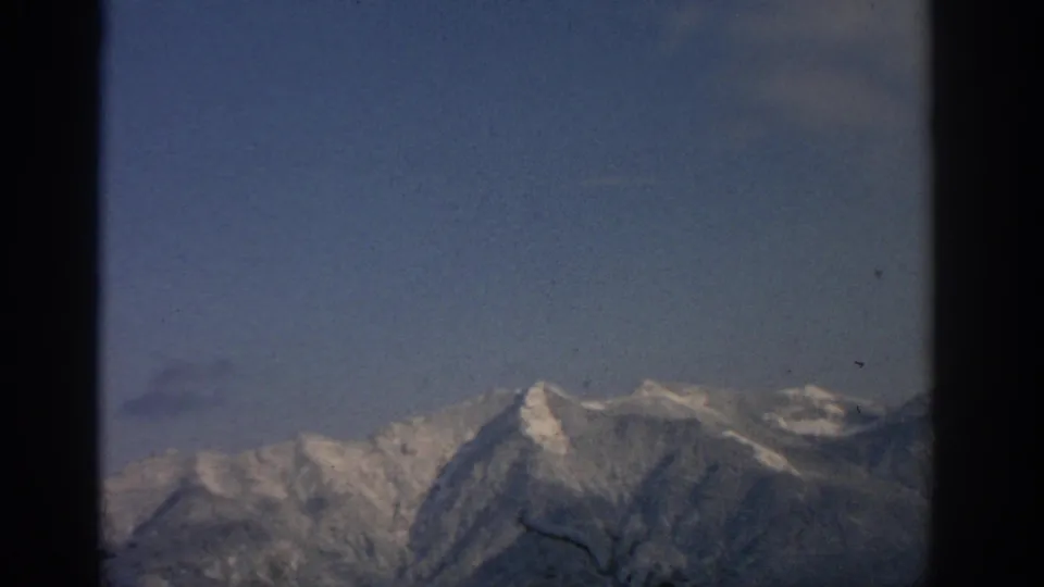 Vintage 8mm frame of snow-covered alpine peaks near Munich, with blue sky, soft haze, and a small dark branch crossing the foreground.