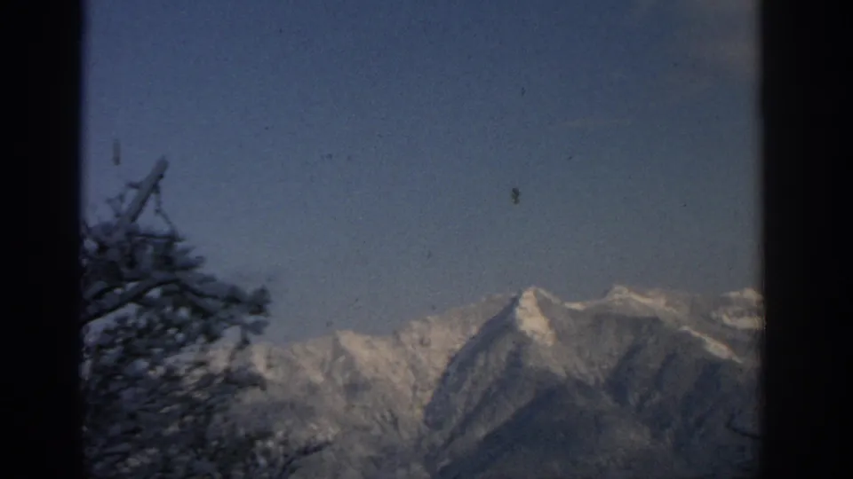 1963 home-movie view near Munich of snow-covered alpine peaks framed by dark evergreen trees against a clear blue sky.