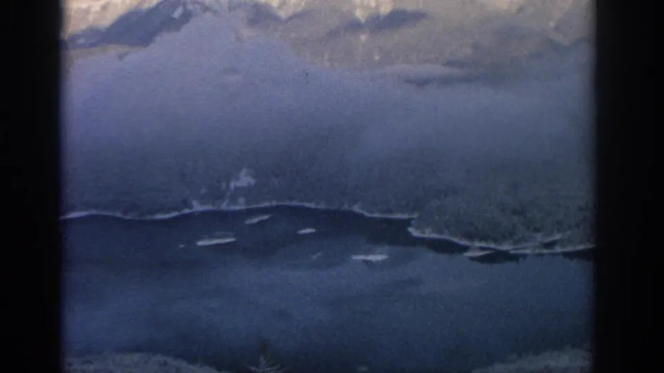 1963 8mm frame showing snow-bright mountain slopes above a dark lake or valley under a pale winter sky.