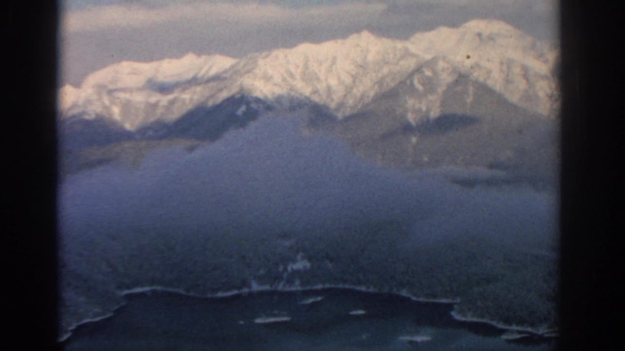1963 8mm aerial view near Munich showing snow-covered mountains, dark forested slopes, haze, and a lake edge below.