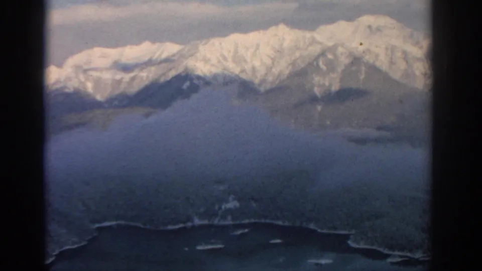 1963 8mm aerial view near Munich showing snow-covered mountains, dark forested slopes, haze, and a lake edge below.