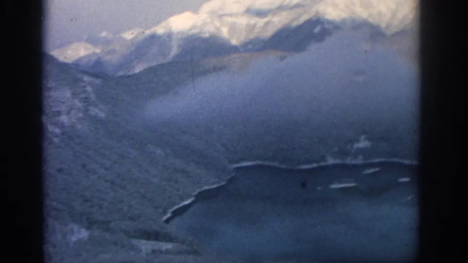 1963 8mm aerial view near Munich looking across a hazy alpine slope toward snow-covered peaks and a blue lake inlet.