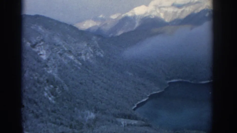 1963 8mm aerial frame near Munich showing a wooded slope, distant snowy mountains, cloud shadow, and the edge of a deep blue lake.