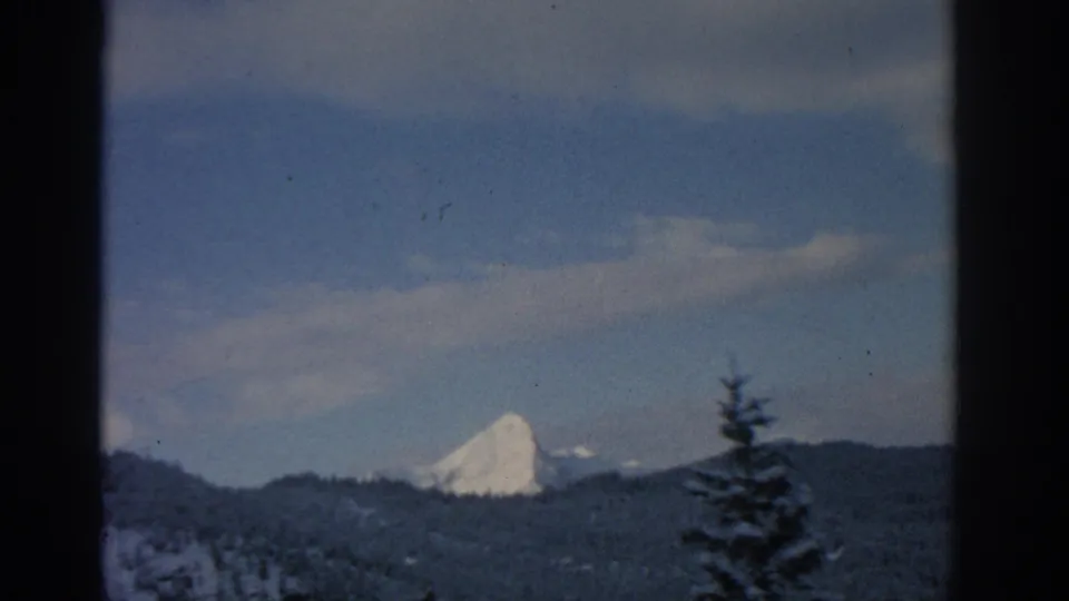1963 Munich-region landscape frame with a snow-capped peak rising beyond dark wooded ridges and sparse clouds.
