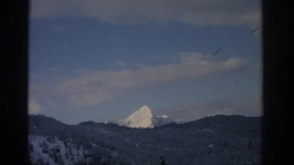 1963 Munich-region 8mm view of a distant snow-capped mountain set behind dark winter ridges beneath pale cloud cover.