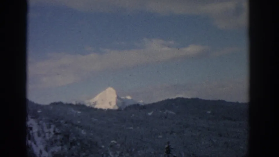 1963 Munich-region frame showing a distant white alpine peak, dark wooded ridges, blue sky, and a foreground evergreen tip.