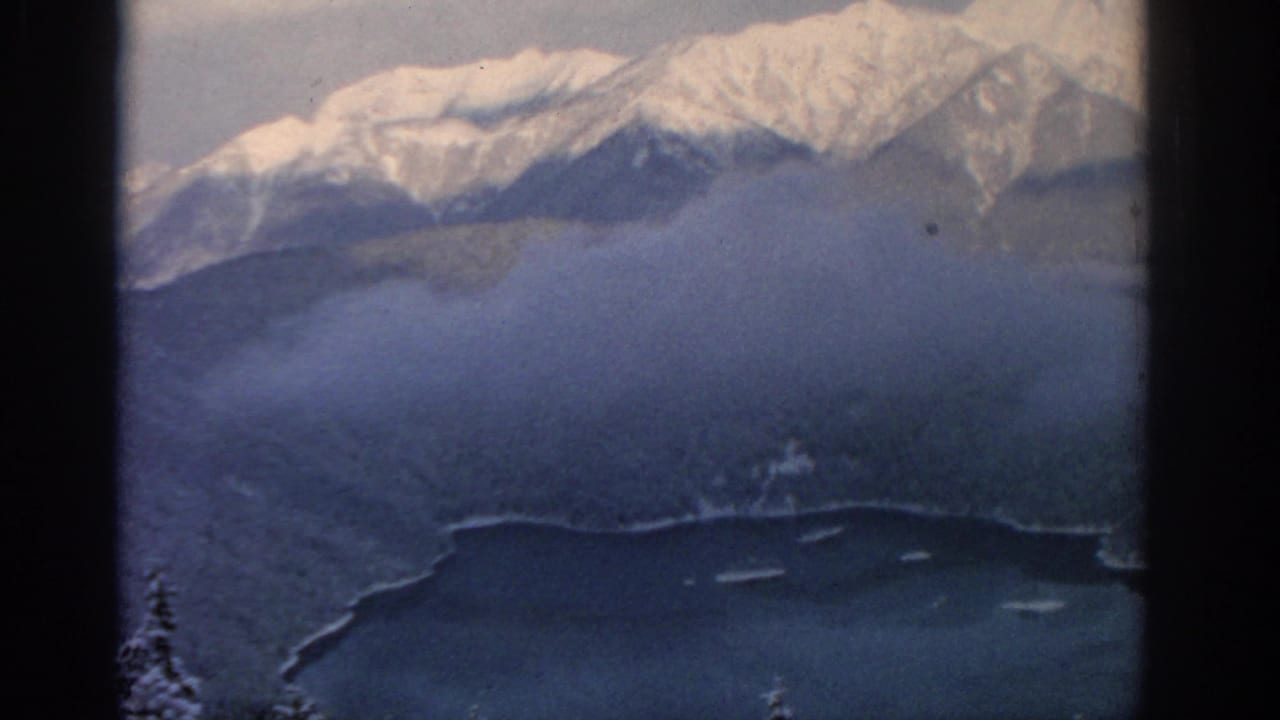 Aerial 8mm frame from 1963 near Munich with snowy alpine peaks, drifting cloud, forest, and lake shoreline.