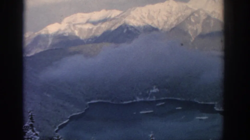 Aerial 8mm frame from 1963 near Munich with snowy alpine peaks, drifting cloud, forest, and lake shoreline.