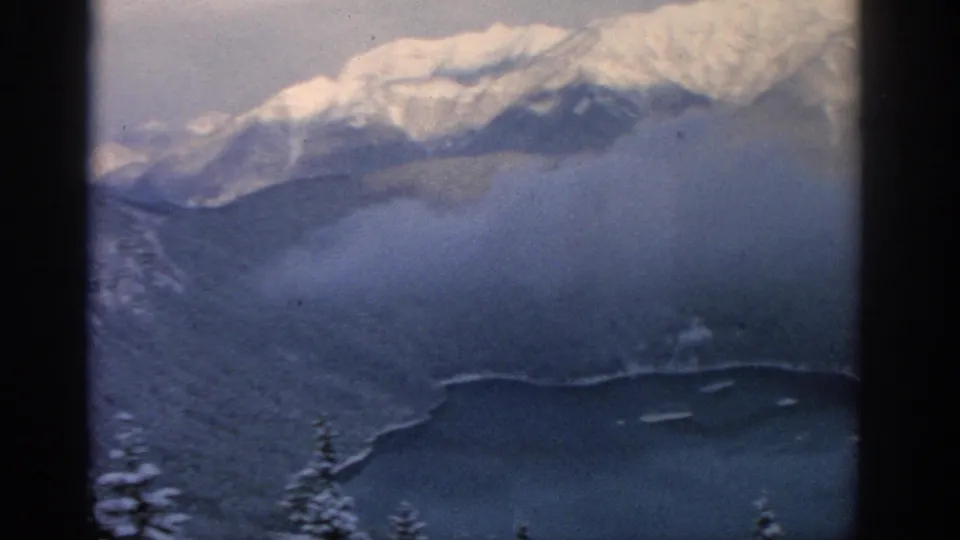 1963 8mm alpine aerial view near Munich, with soft snow peaks above clouded forest and a dark lake below.