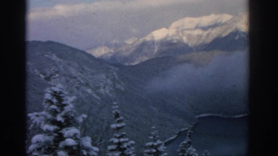 1963 8mm view near Munich showing foreground snowy trees, misty alpine slopes, distant peaks, and lake edge.