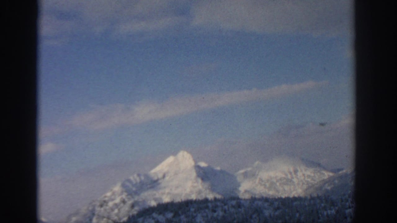 1963 8mm home-movie frame of distant snowy mountain peaks near Munich, Germany, beneath a pale blue sky and thin clouds.