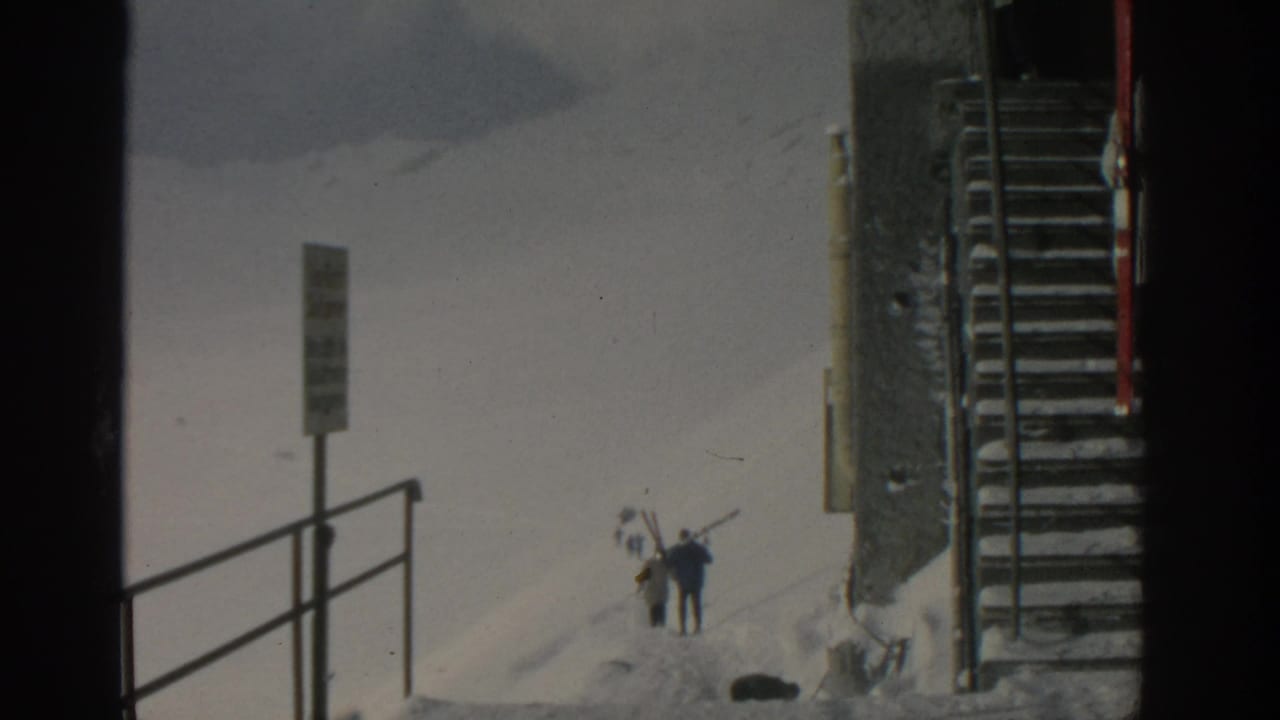 Skiers carrying skis across a snowy slope near a signpost and metal stairway.
