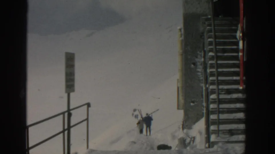 At a snowy ski resort area near Munich, distant skiers carry skis beside a signpost, railing, and exterior metal stairs.