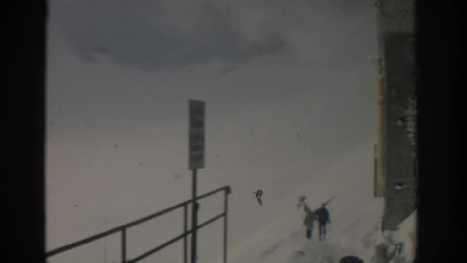 Handheld 8mm view of a Munich-area ski slope with distant skiers, metal railing, signpost, and building wall in deep snow.