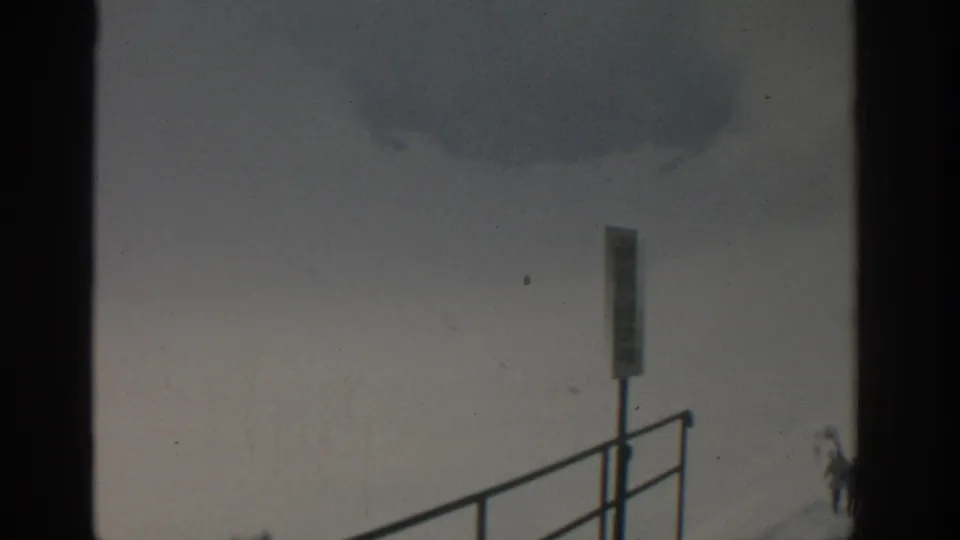 Late frame from the 1963 Munich ski-resort footage showing a signpost, railing, deep snow, and a partial distant skier at frame edge.