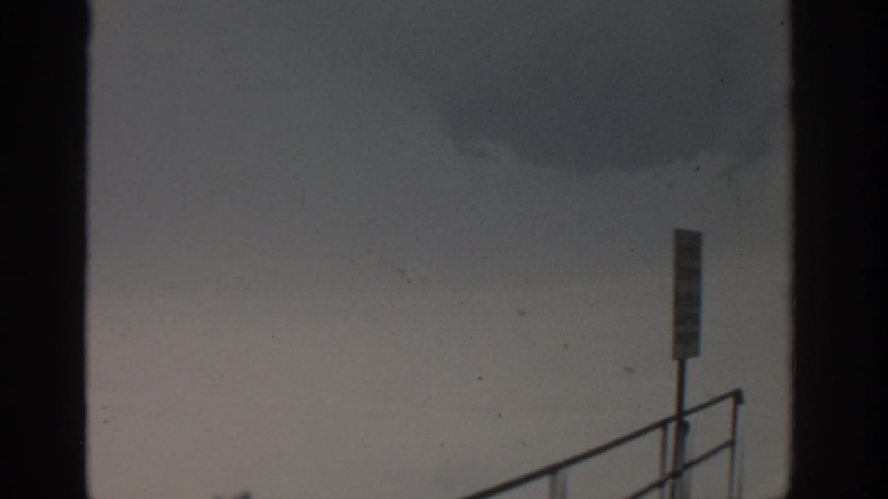 Overcast sky with a faint smoke plume, metal railing, and narrow signpost near the lower frame.