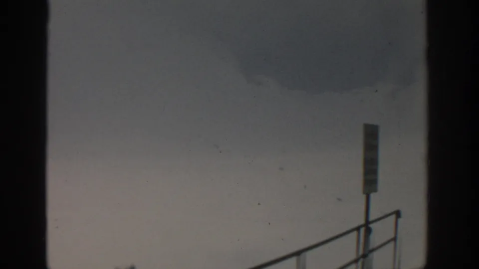 1963 Munich 8mm frame showing hazy sky, faint dark smoke, a railing, and a small signpost from an elevated viewpoint.