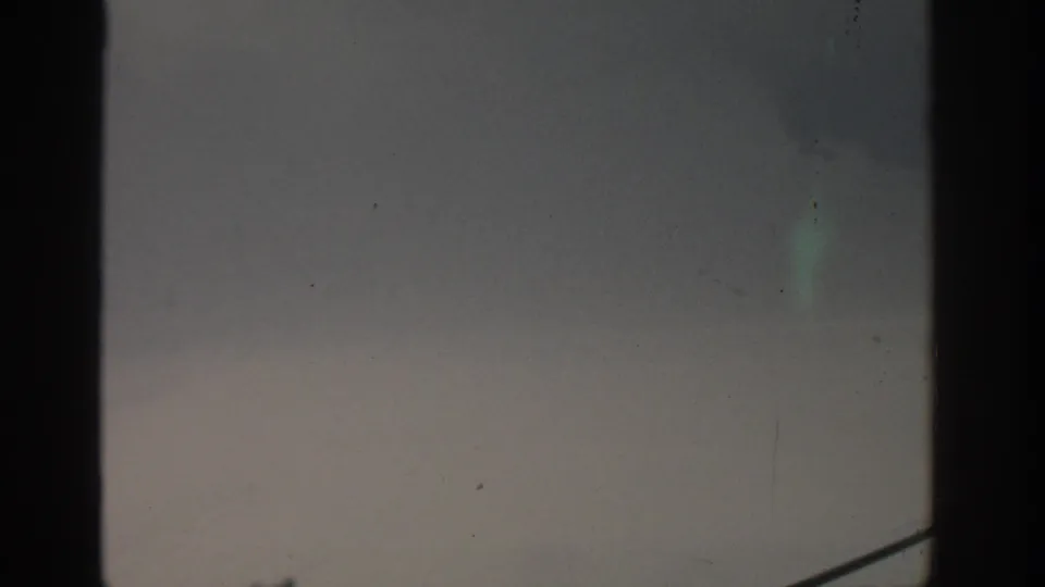 1963 Munich 8mm frame with subdued gray sky, faint drifting smoke, and a partial railing at the lower right.