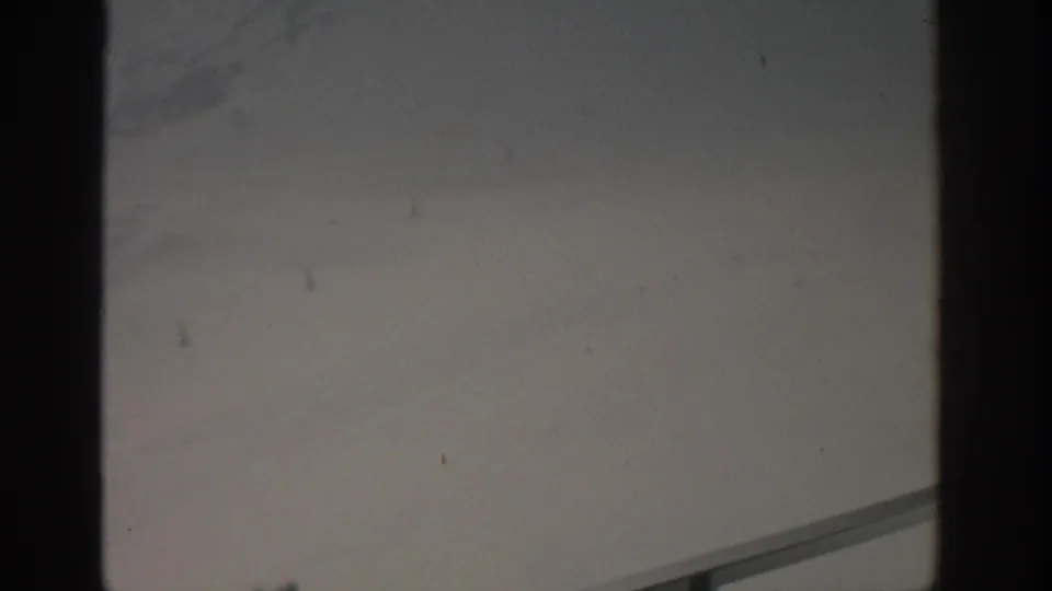 1963 Munich home-movie frame showing a washed gray exterior view through dark window mullions, with faint distant marks and visible film wear.