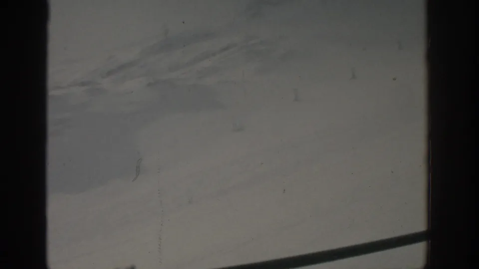 1963 Munich home-movie frame with a pale gray outdoor view, dark window edges, faint distant forms, scratches, and a diagonal rail near the bottom.