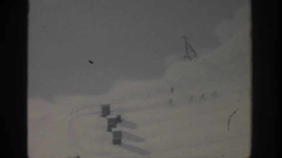 1963 8mm frame of a Munich-area snowy slope with lift structures, faint tracks, and a distant skier on the hillside.