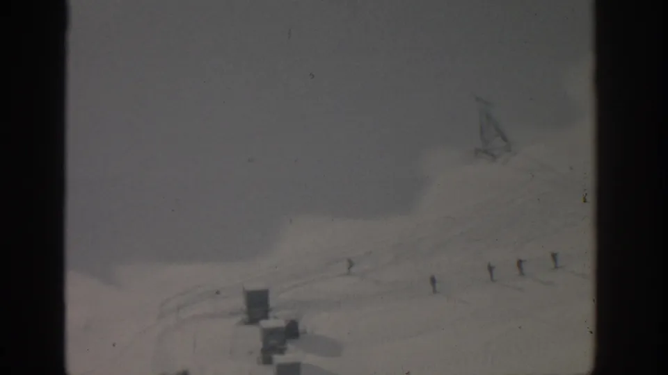 Munich-area 1963 home-movie frame showing a small skier on a broad snow slope beside simple ski-lift structures.