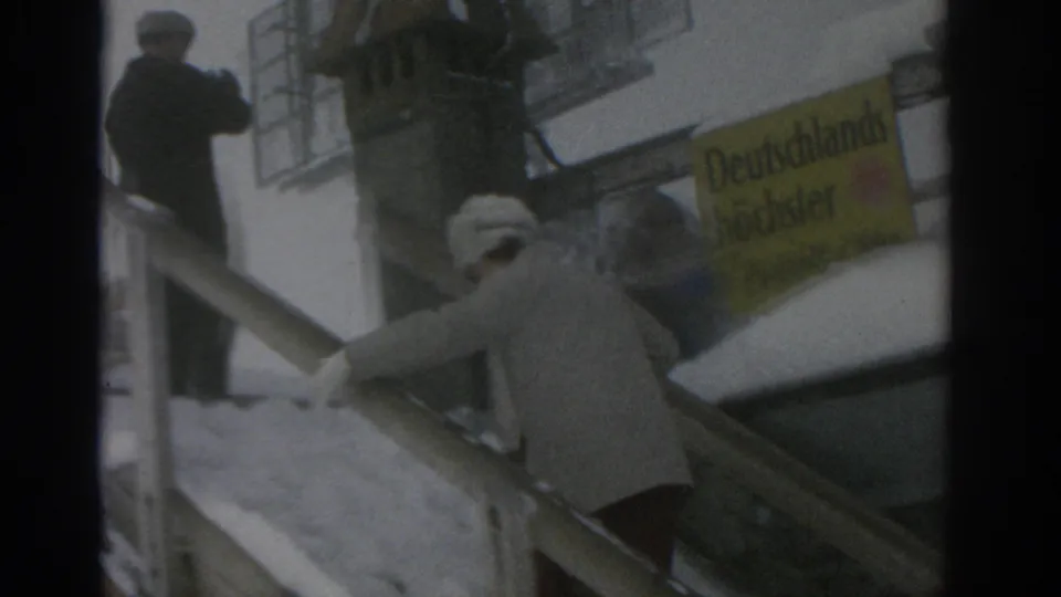 In the supplied 1963 Munich context, adults in winter coats move up snow-covered stairs near a yellow German sign.