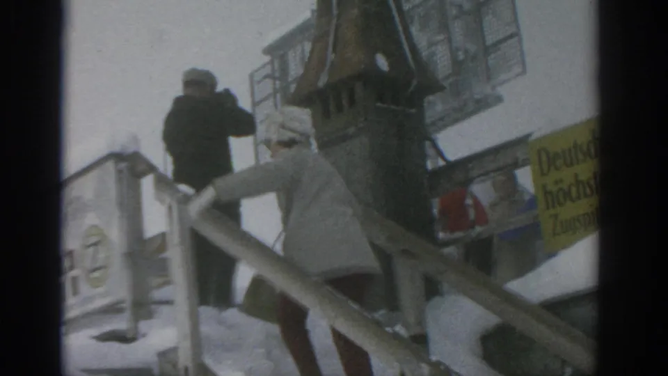 A woman in a light winter coat ascends snowy stairs while another bundled adult stands or climbs ahead near a yellow sign.