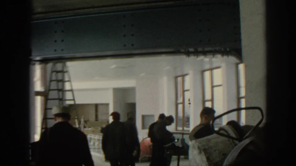 Munich 1963 8mm frame showing people in coats and caps moving inside a construction interior with ladder, windows, and exposed beams.