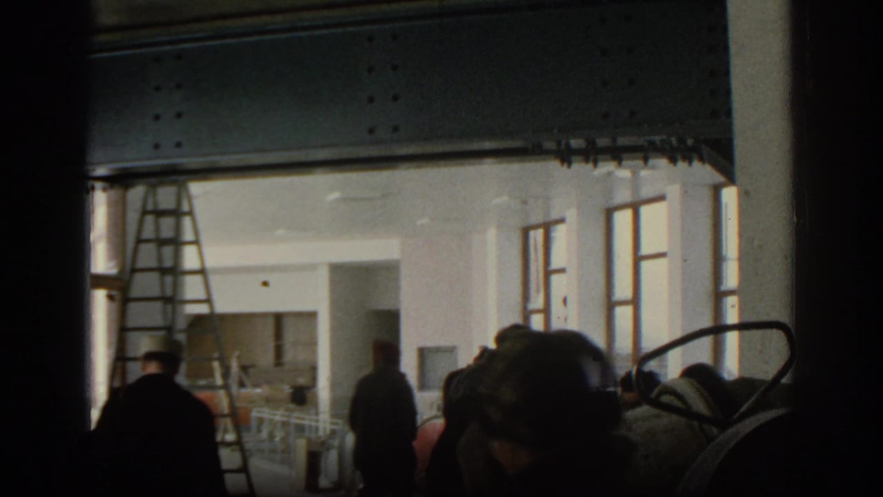 Figures walk inside a bright unfinished room with ladder, windows, beams, and equipment in the foreground.