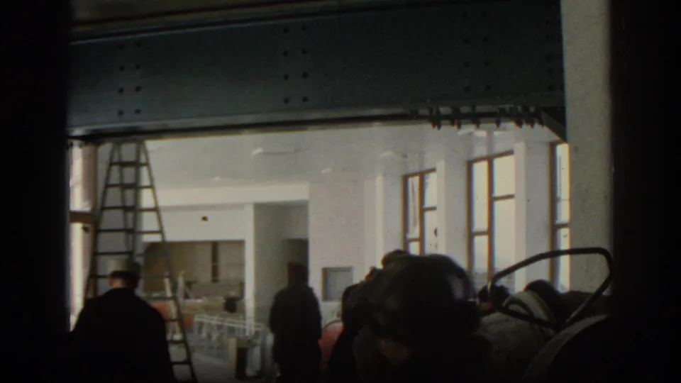 Munich 1963 frame of people crossing a construction-site interior, with a ladder near tall windows and dark equipment partly blocking the view.