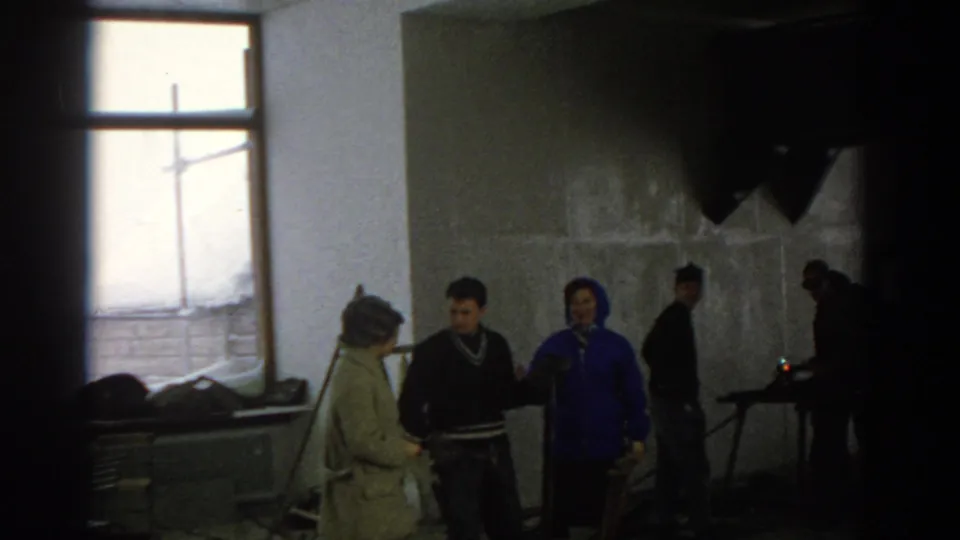 Munich 1963 8mm frame showing a woman in a green coat speaking with others in an unfinished room with concrete walls and window light.