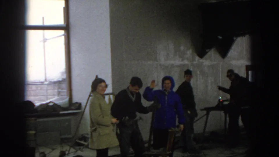 Munich 1963 frame of people conversing inside a construction interior, with unfinished concrete walls, a window, and work equipment nearby.