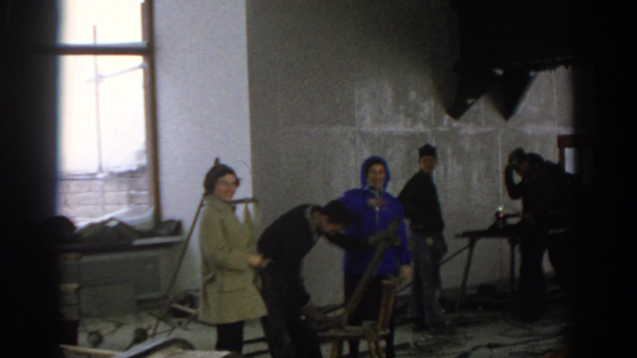 Munich 1963 home-movie frame of adults in coats and work clothes gathered in an unfinished room as a man works near a chair.