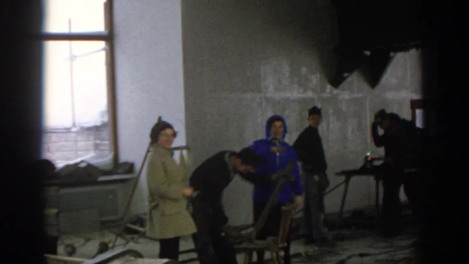 Munich 1963 home-movie frame of adults in coats and work clothes gathered in an unfinished room as a man works near a chair.